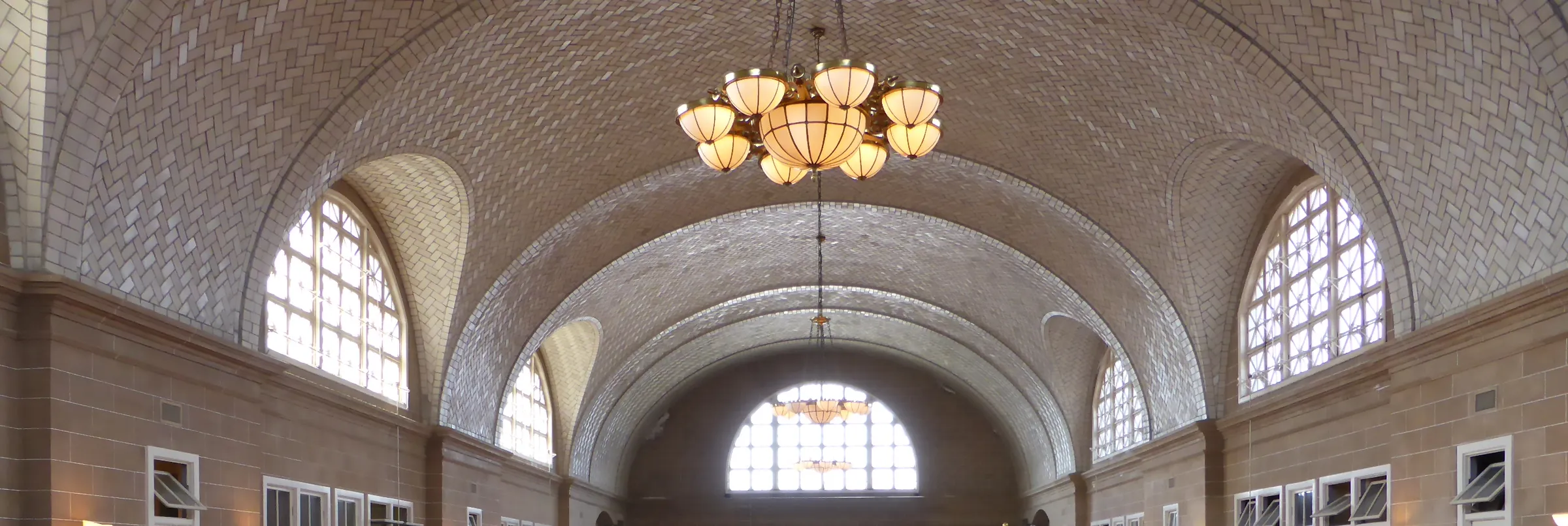 The Guastavino tile vaulted ceiling of the Ellis Island Immigration Museum hall, with arched windows and a hanging chandelier.